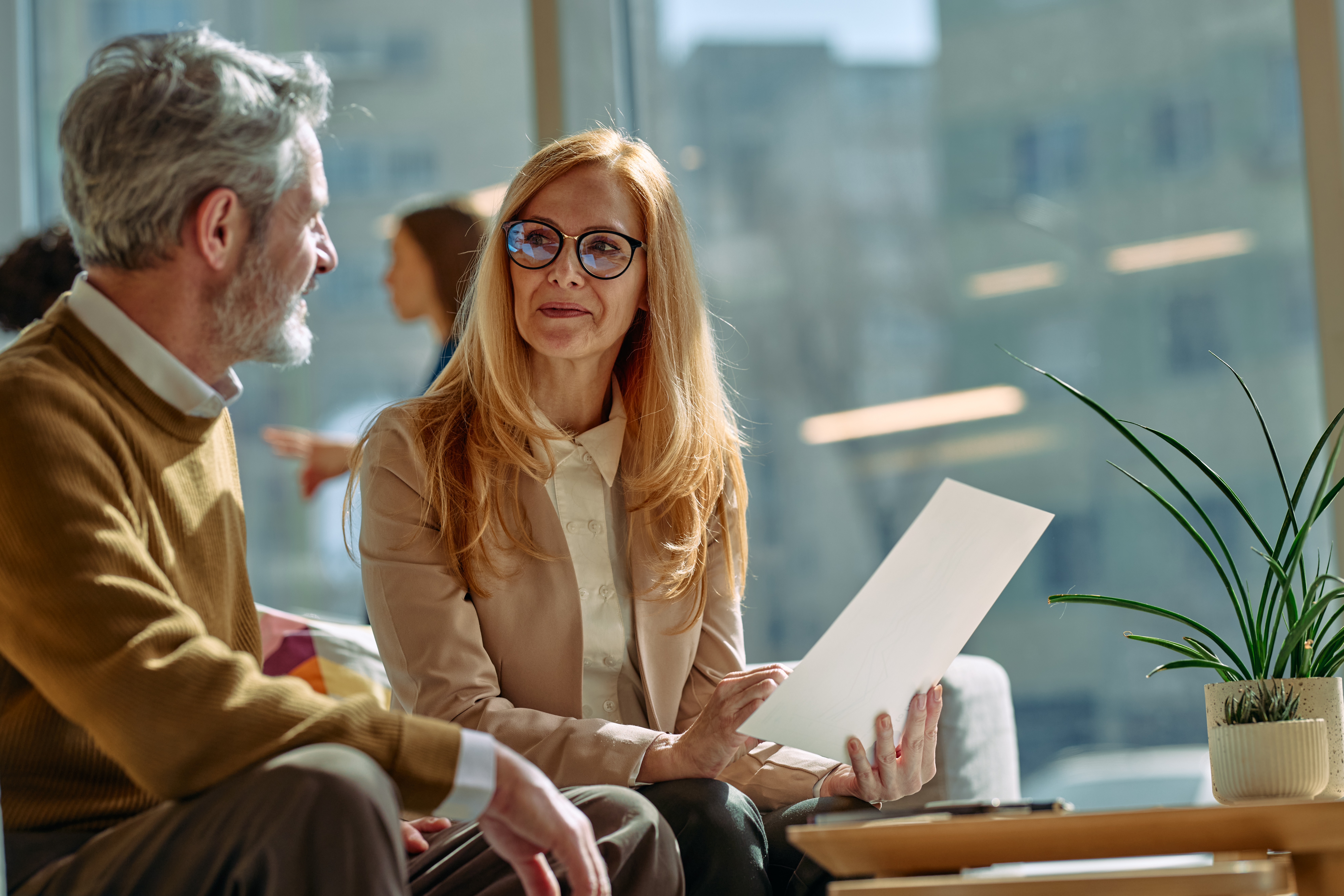 Two mature business people sitting on a sofa, discussing a document during a relaxed team meeting in a modern office